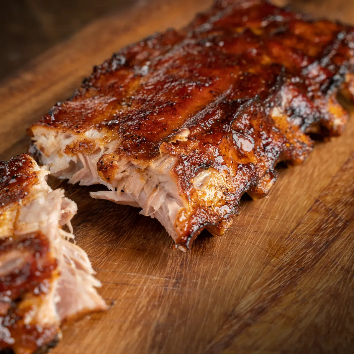 A close-up of a signature Mr MIKES Steakhouse plate featuring a glazed rack of ribs on a wooden board, with a piece pulled away to reveal the tender, juicy meat inside.