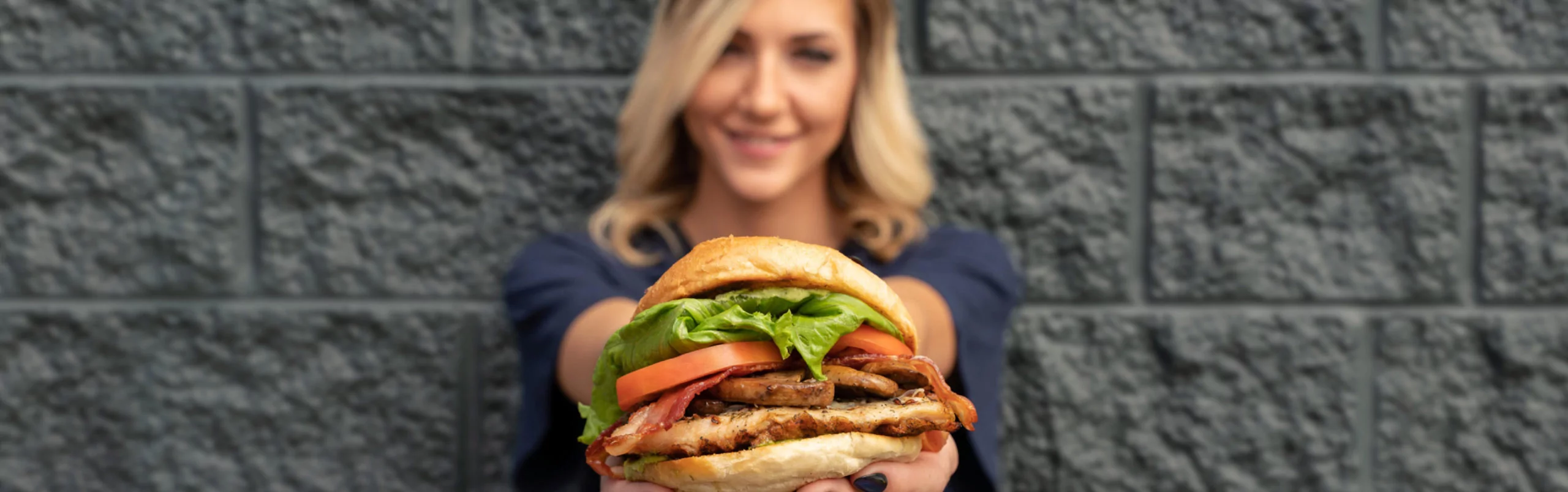 A woman holds out a "Mikeburger" from Mr MIKES Steakhouse, stacked with lettuce, tomato, bacon, and a grilled patty.