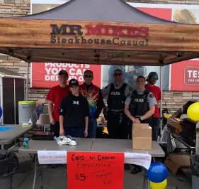 Volunteers and police officers stand under a Mr MIKES Steakhouse Casual tent at a community fundraising event, with a sign promoting 