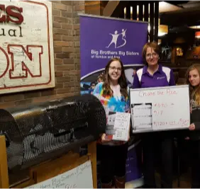 A group at a Big Brothers Big Sisters fundraiser at Mr MIKES Steakhouse, standing by a raffle drum and donation board.