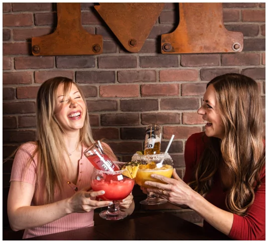 Two women share a laugh while enjoying large, colorful drinks at Mr MIKES Steakhouse, with the restaurant’s rustic logo mounted on a brick wall in the background.