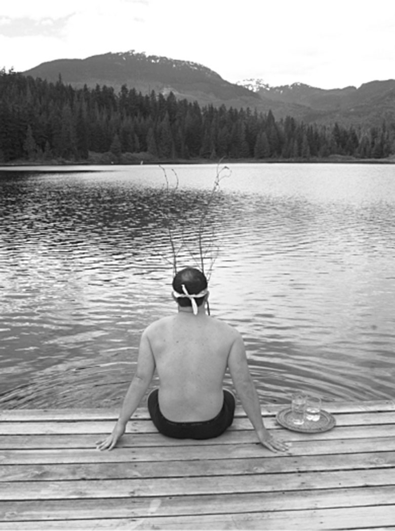 Person sitting on a dock by a peaceful lake surrounded by mountains and trees.